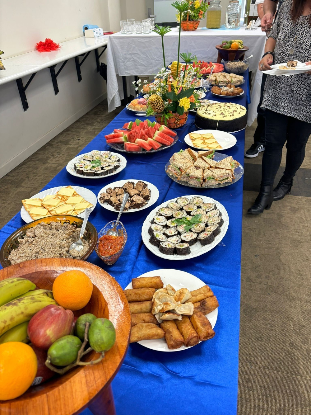 A buffet table is set with a variety of dishes, including sushi, fruit, pastries, and desserts on a blue tablecloth.