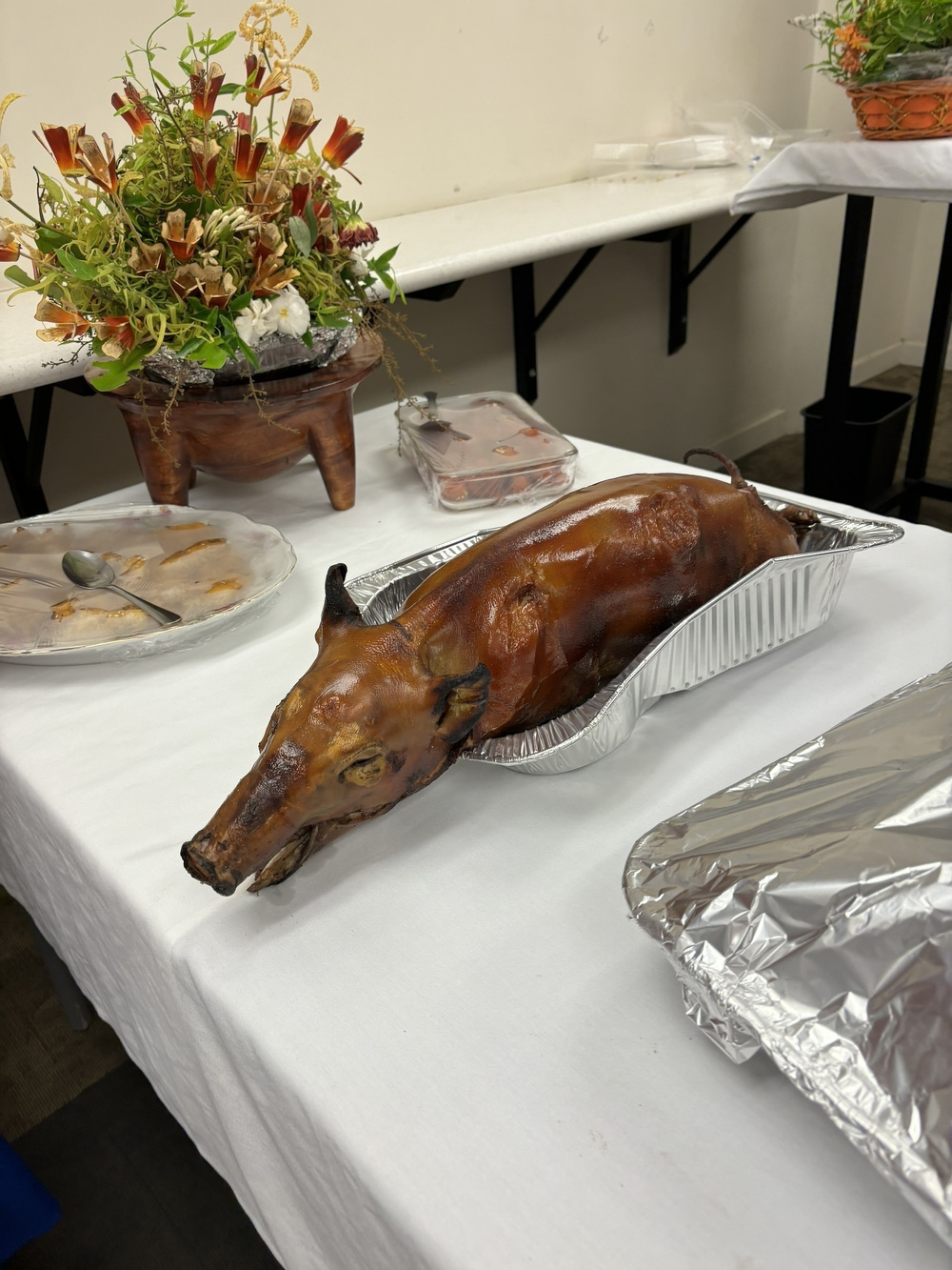 A roasted pig is presented on a table alongside various dishes and a decorative floral arrangement.