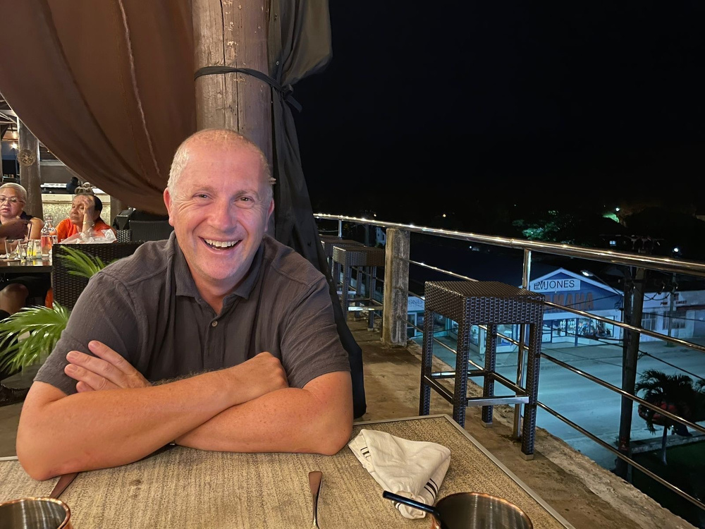 A smiling person is seated at an outdoor restaurant table at night.