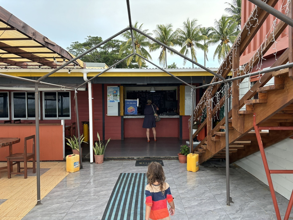 A child walks towards a small outdoor cafe or kiosk underneath a canopy, with a palm tree-lined sky in the background.