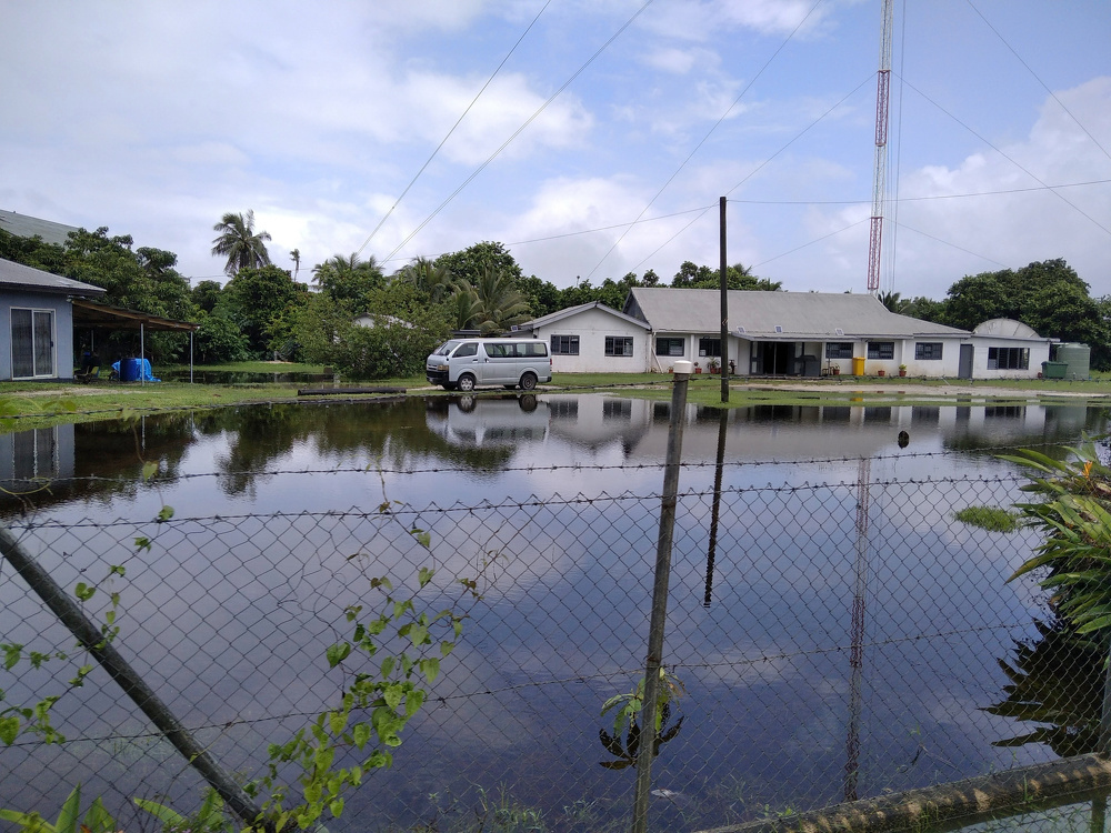 A fenced yard is flooded with water, reflecting a van, houses, and an overcast sky.