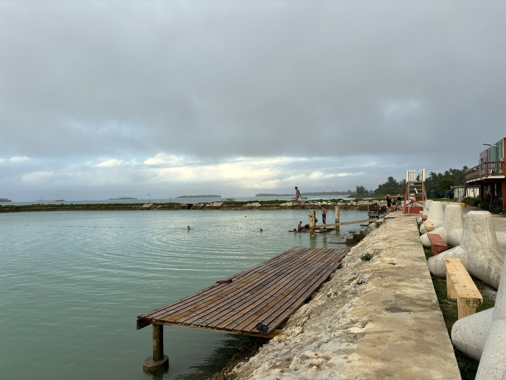 A wooden pier extends into a calm body of water where several people are swimming, with a rocky shoreline and cloudy sky in the background.