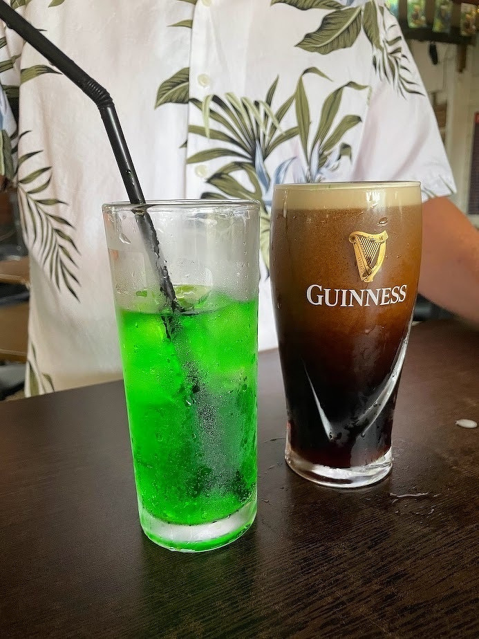 Two drinks, one a green beverage in a glass with a straw and the other a pint of Guinness, are placed on a table in front of a person wearing a white shirt with a leaf pattern.