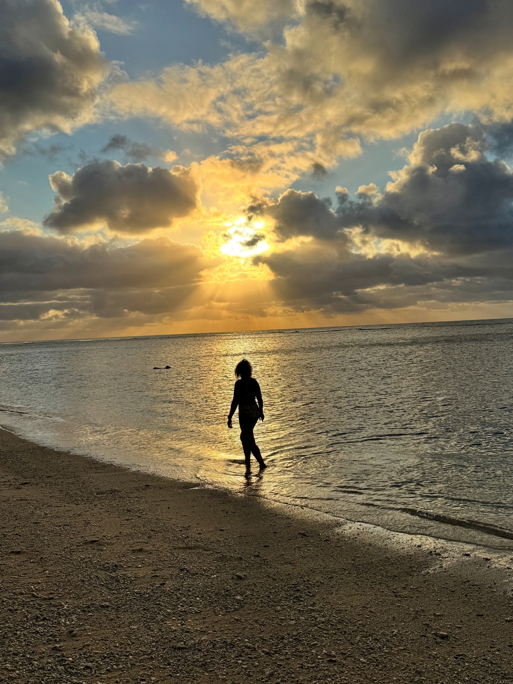 A person walks along a tranquil beach with a dramatic sunset casting light through the clouds.