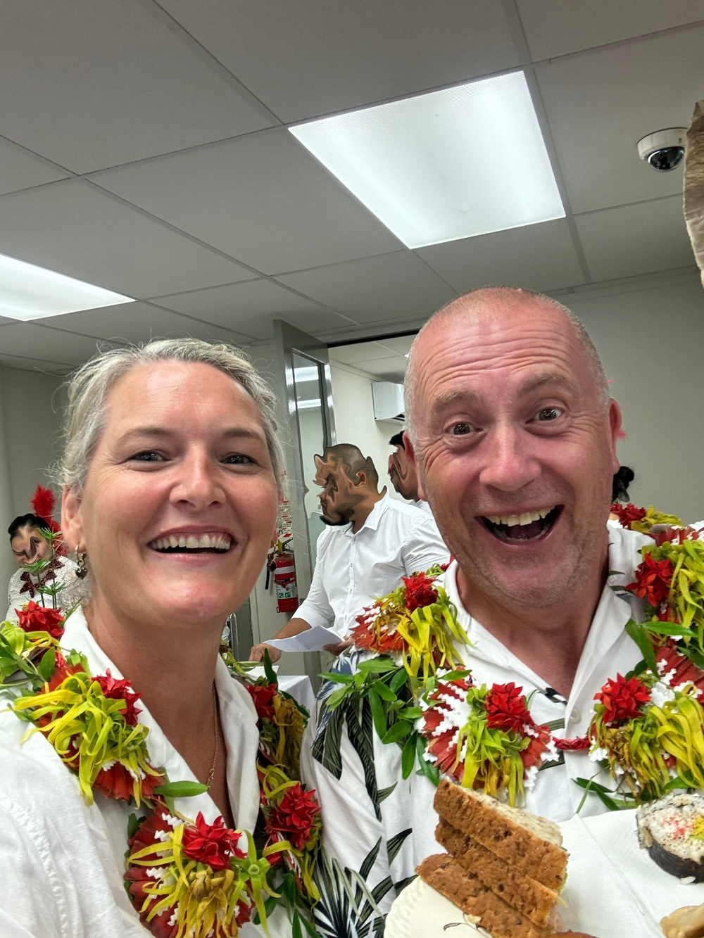 A smiling man and woman wearing Hawaiian leis take a cheerful selfie indoors with snacks in hand.