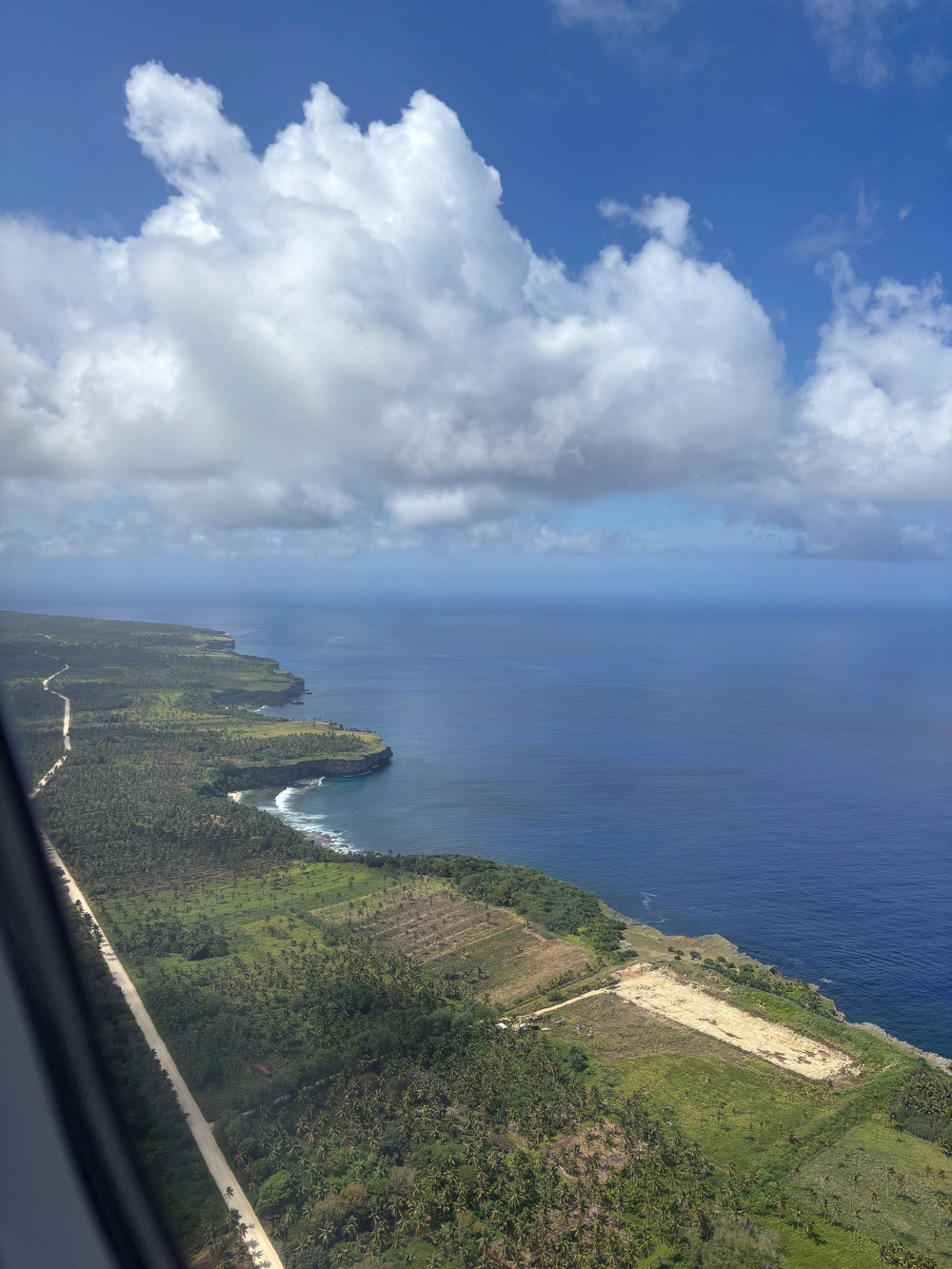 Aerial view of a coastal landscape featuring lush greenery, sprawling fields, a winding road, and expansive ocean beneath a partly cloudy sky.