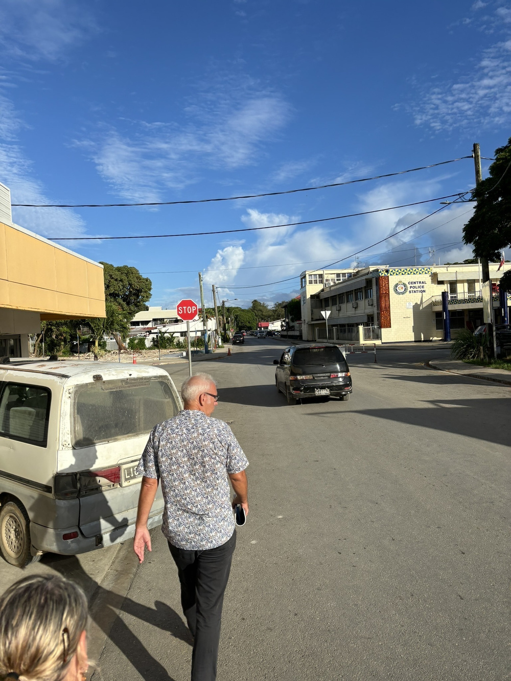A person walks down a quiet street with buildings, parked vehicles, and a stop sign visible under a partly cloudy sky.