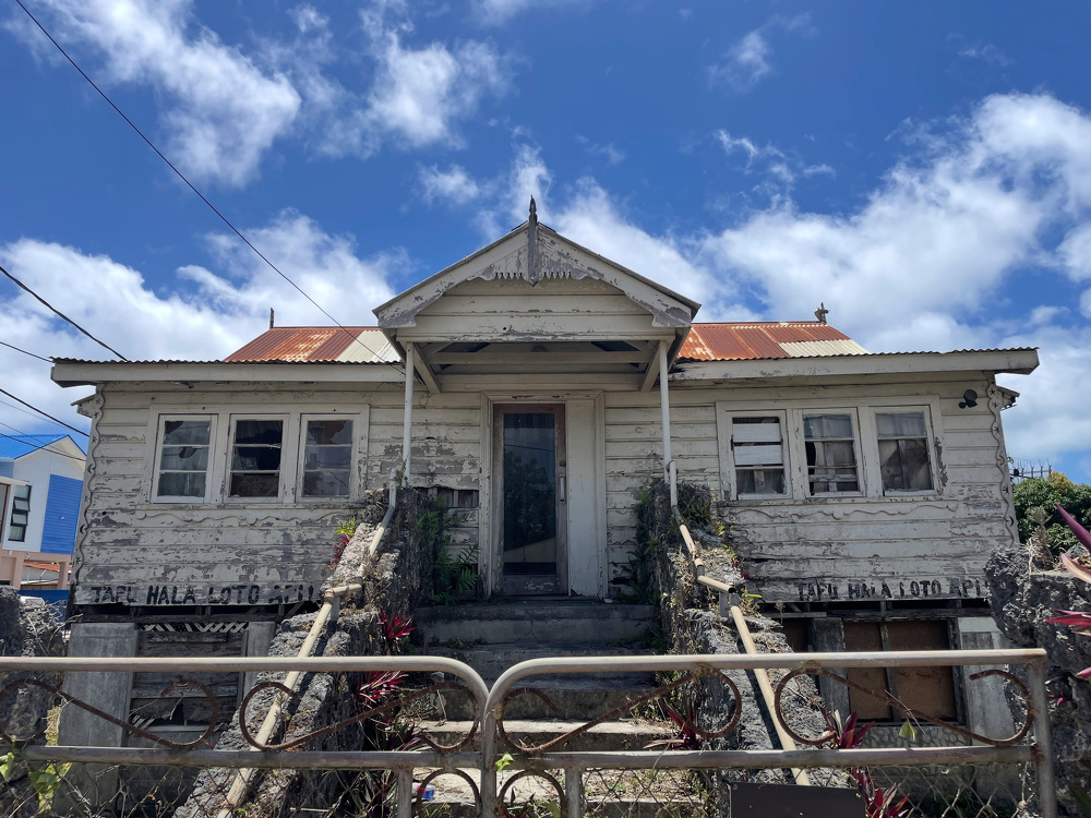 A weathered, abandoned house with a rusted roof and peeling paint stands behind a gated entrance, set against a partly cloudy sky.