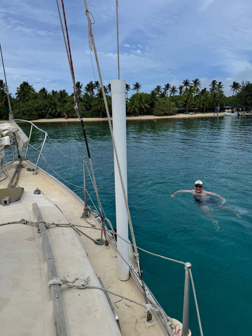 A person wearing a hat is swimming near a sailboat in clear blue water, with a tropical island and palm trees in the background.