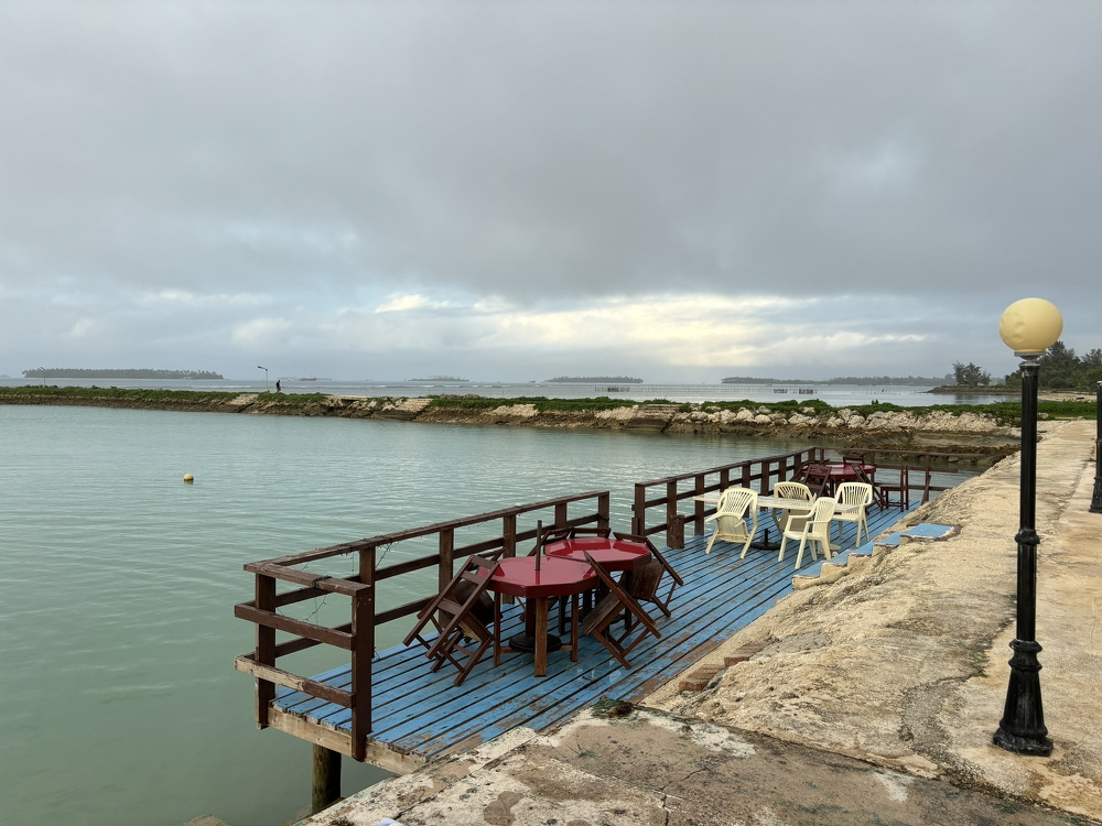 A small wooden deck with tables and chairs overlooks a calm body of water under a cloudy sky.
