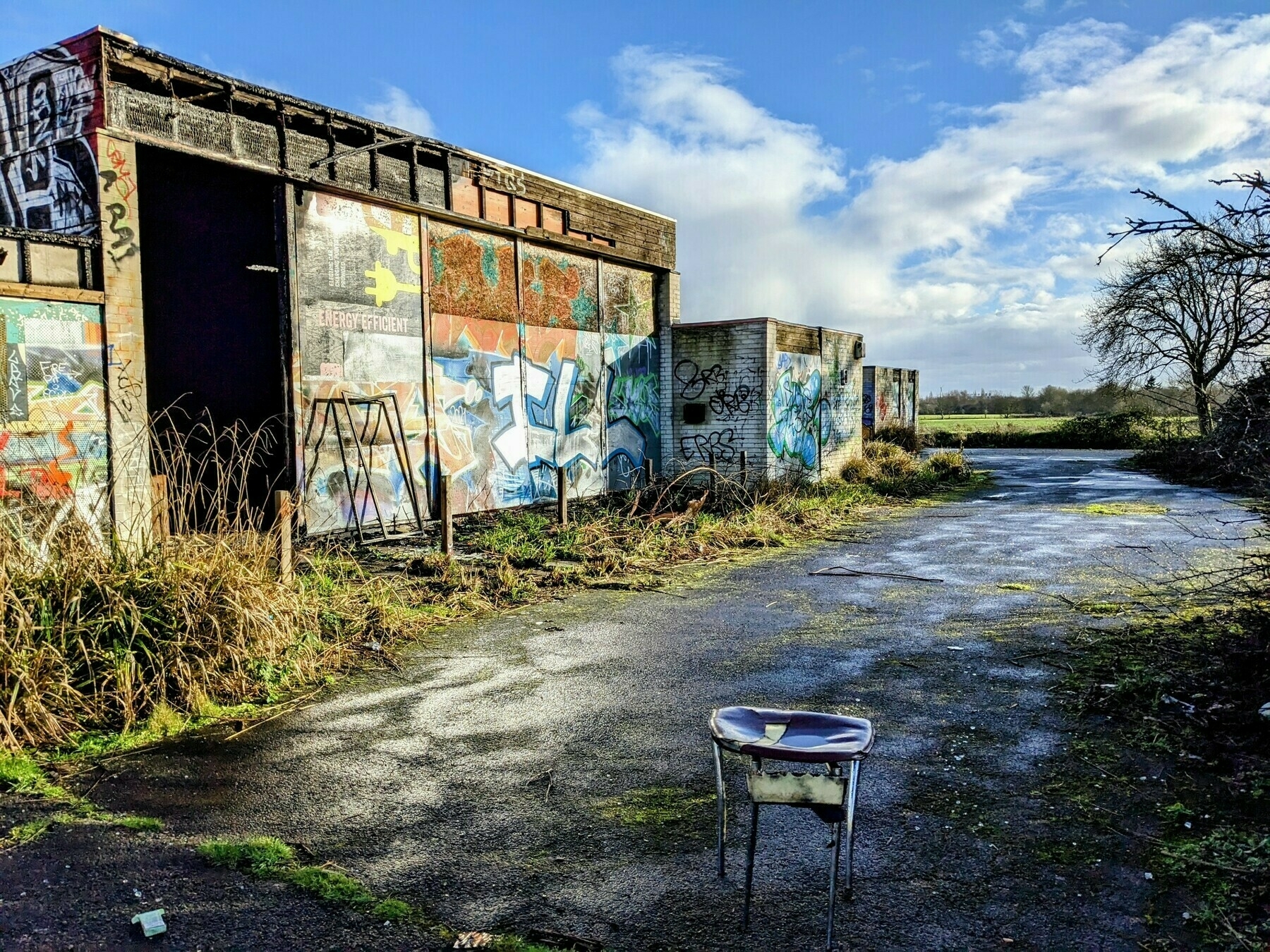 An abandoned, graffiti-covered building sits beside a wet pathway, under a bright blue sky with scattered clouds.