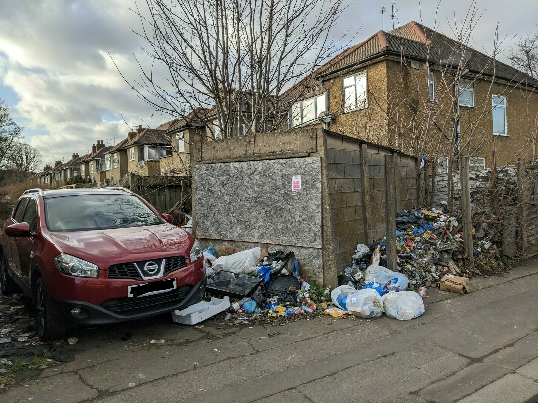 Photo of car parked in alleyway surrounded by fly-tipped and dumped rubbish. Faded sign on the wall behind says: NO TIPPING OR DUMPING