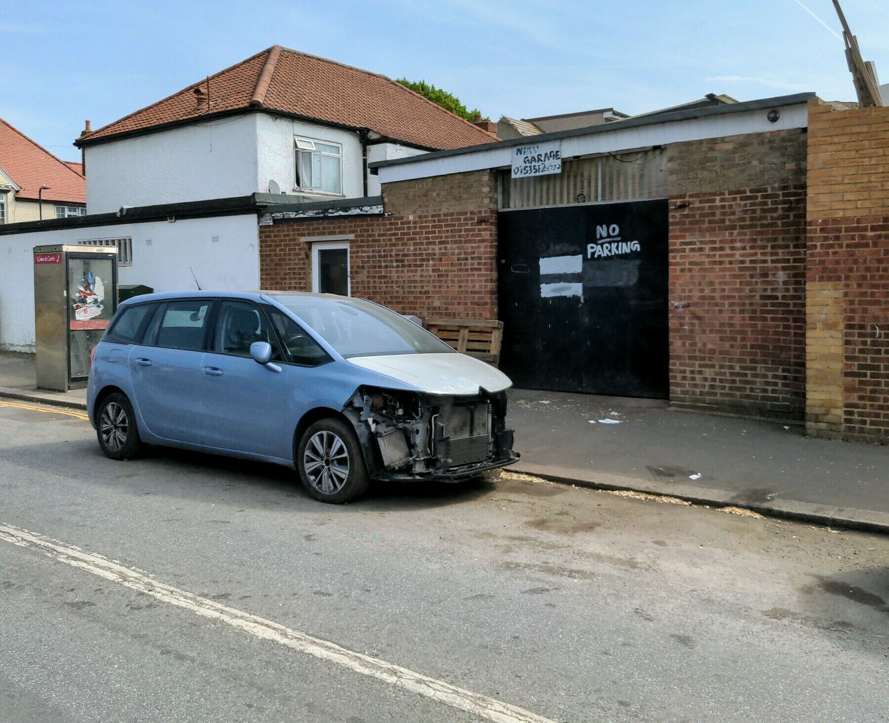 Photo of a car parked outside a garage displaying a handwritten "No Parking" sign in Southall Green. The front of the car is missing.