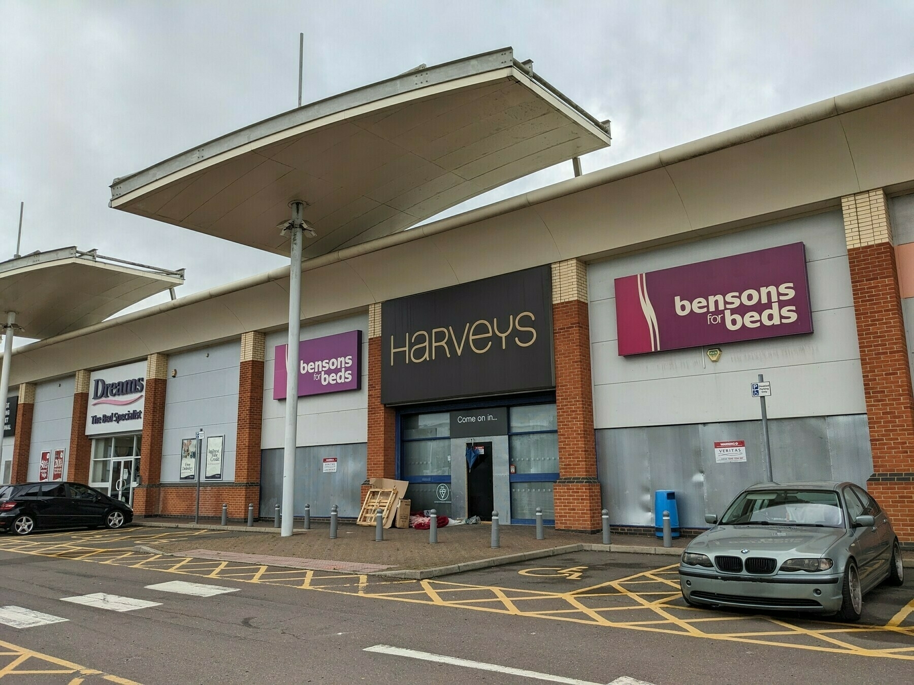 A homeless person's makeshift bed and shelter outside a trading estate premises with apposite signs above the closed doors: Harveys "Come on in..."; Bensons "for beds"; and Dreams "The Bed Specialist".