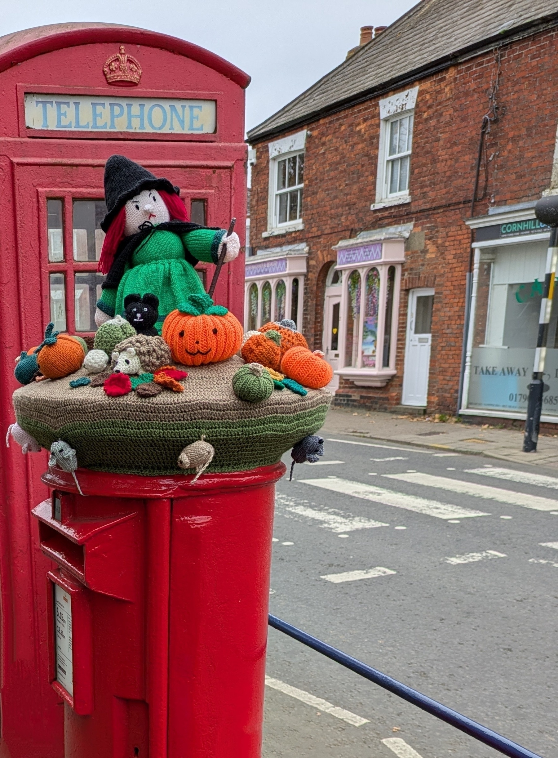 A red post box in front of a red telephone booth features a whimsical knitted display with a witch and various pumpkins on top, and mice below.