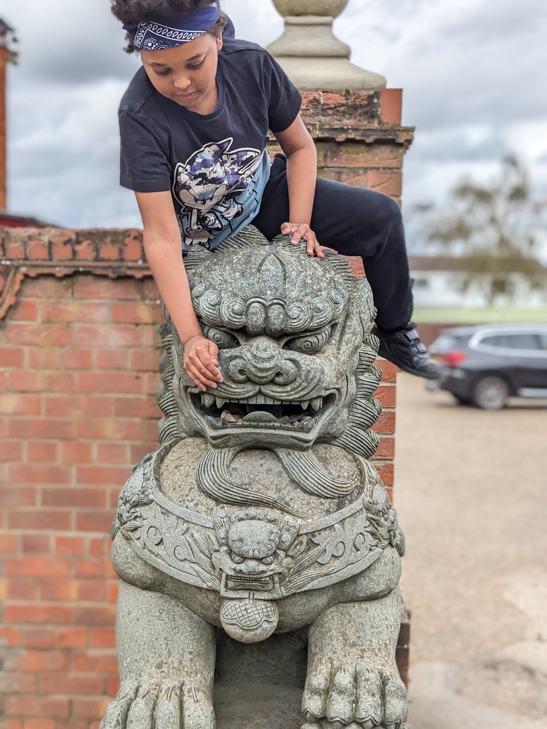 A person wearing a bandana and graphic t-shirt is playfully sitting on a stone lion statue.