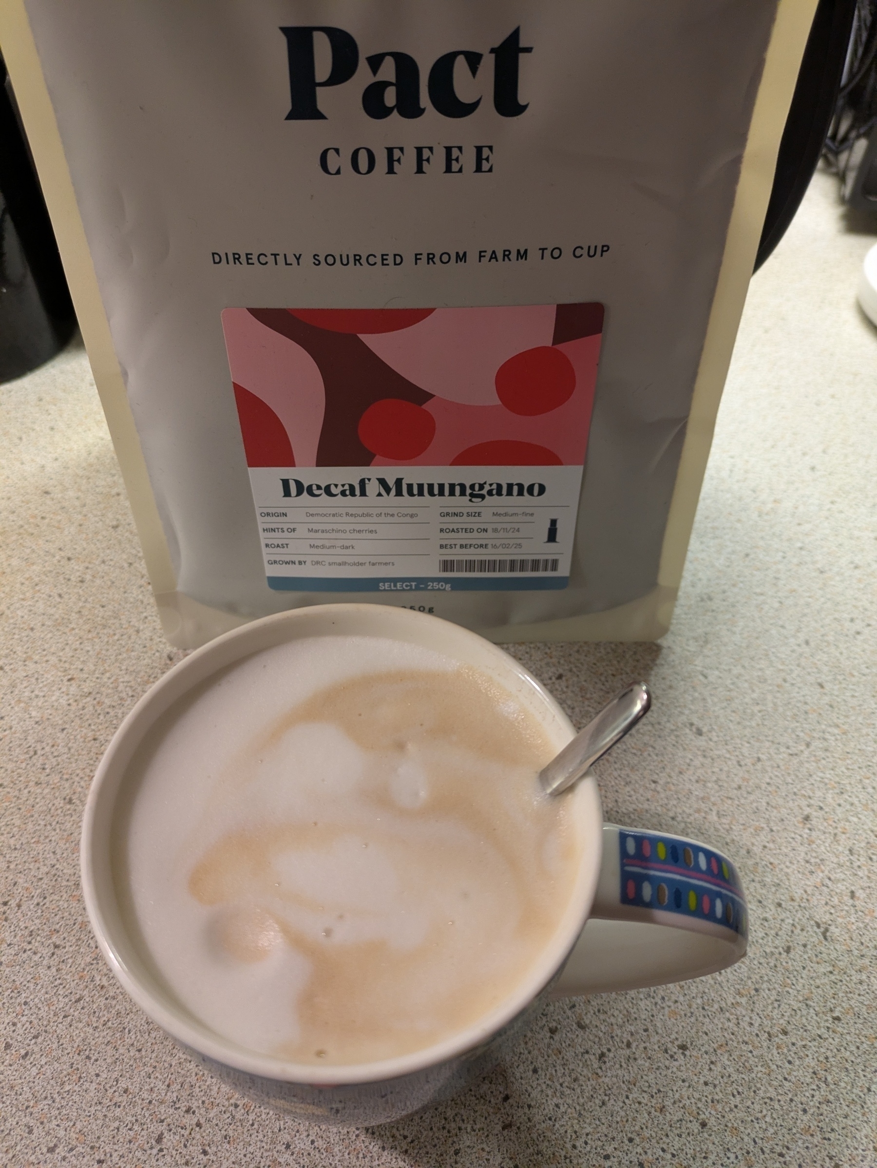 A cup of coffee with frothy milk sits in front of a package of Pact Coffee labeled "Decaf Muungano."