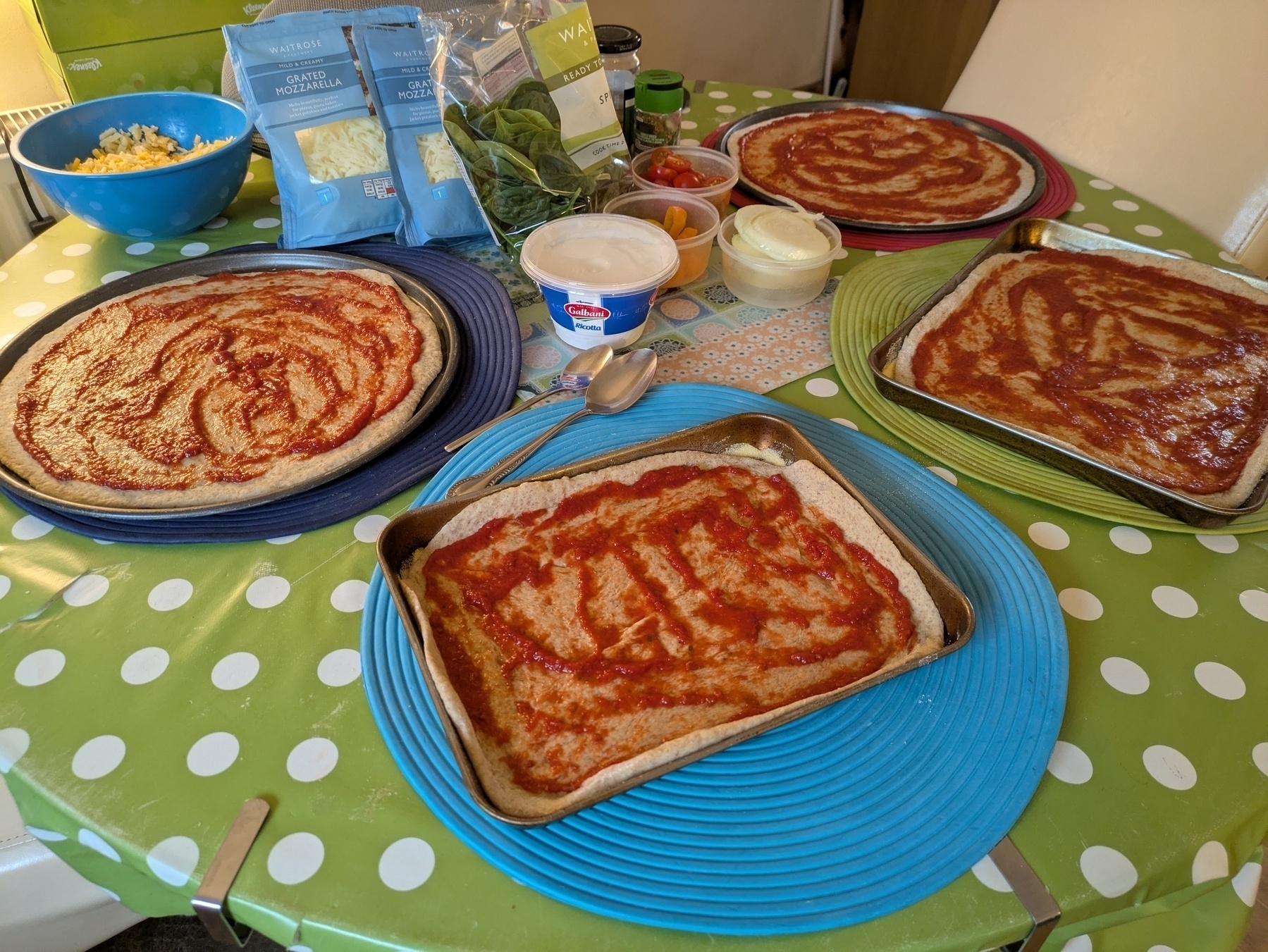 Pizza doughs with tomato sauce are prepared on plates and trays, surrounded by various toppings and ingredients on a polka dot tablecloth.