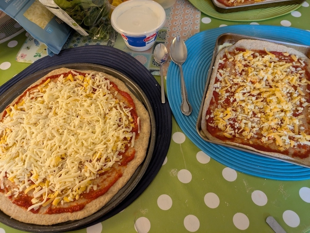 Two uncooked pizzas with cheese and sauce are on a polka dot tablecloth, surrounded by kitchen items.