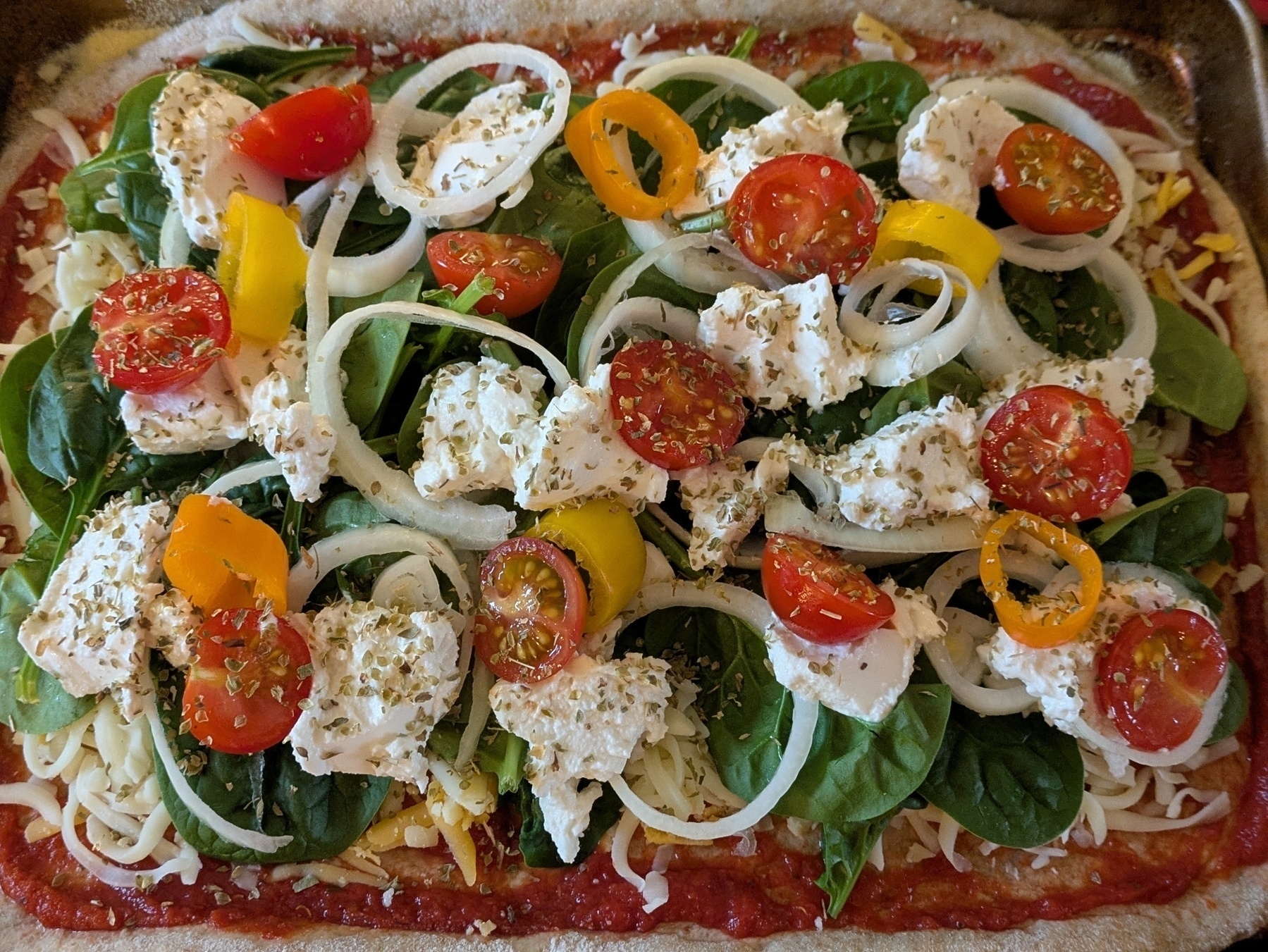 A colorful homemade pizza is topped with fresh spinach, sliced onions, cherry tomatoes, yellow bell peppers, and dollops of ricotta cheese.