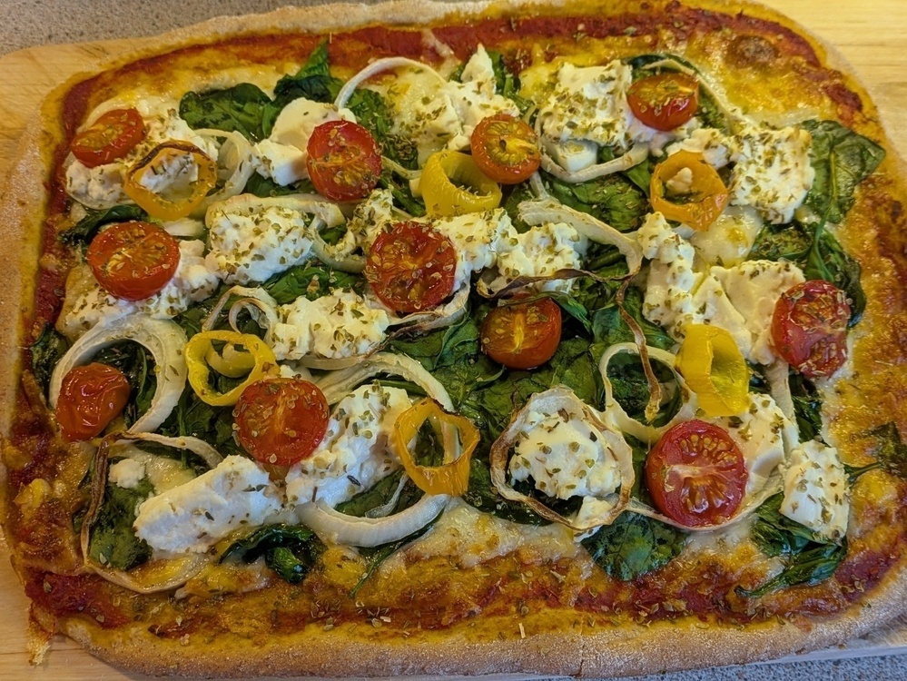 A homemade pizza topped with spinach, cherry tomatoes, onion slices, and dollops of cheese is displayed on a wooden board.