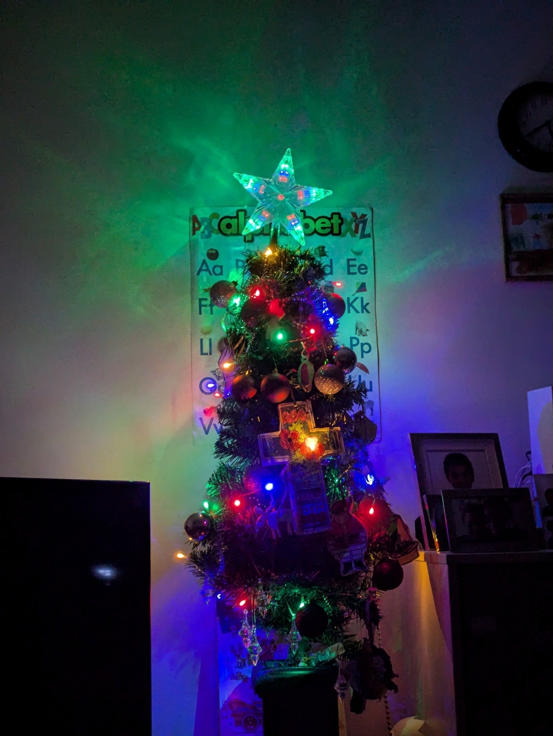 A decorated Christmas tree is illuminated with colorful lights and topped with a glowing star, set against a background that includes an alphabet chart and a few framed photos.