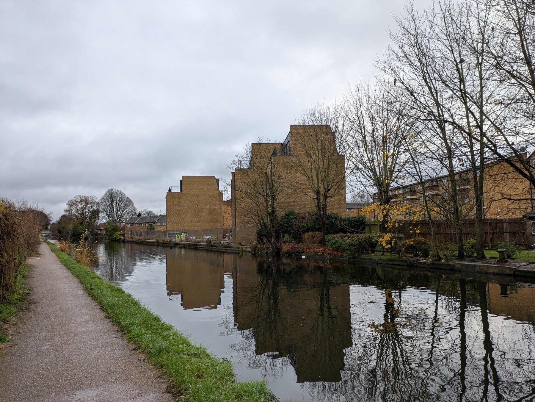 A canal lined with bare trees runs alongside a path and unique, blocky buildings on a cloudy day.