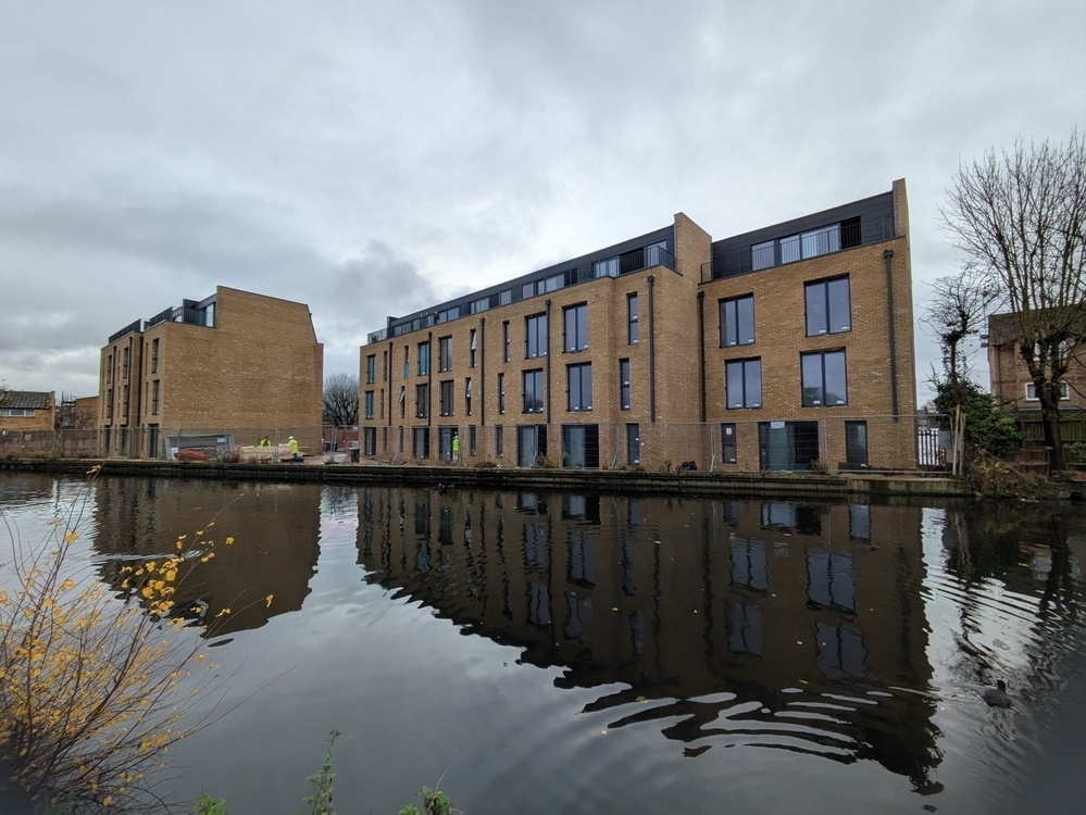 Modern, multi-story apartment buildings are reflected in a calm body of water on a cloudy day.