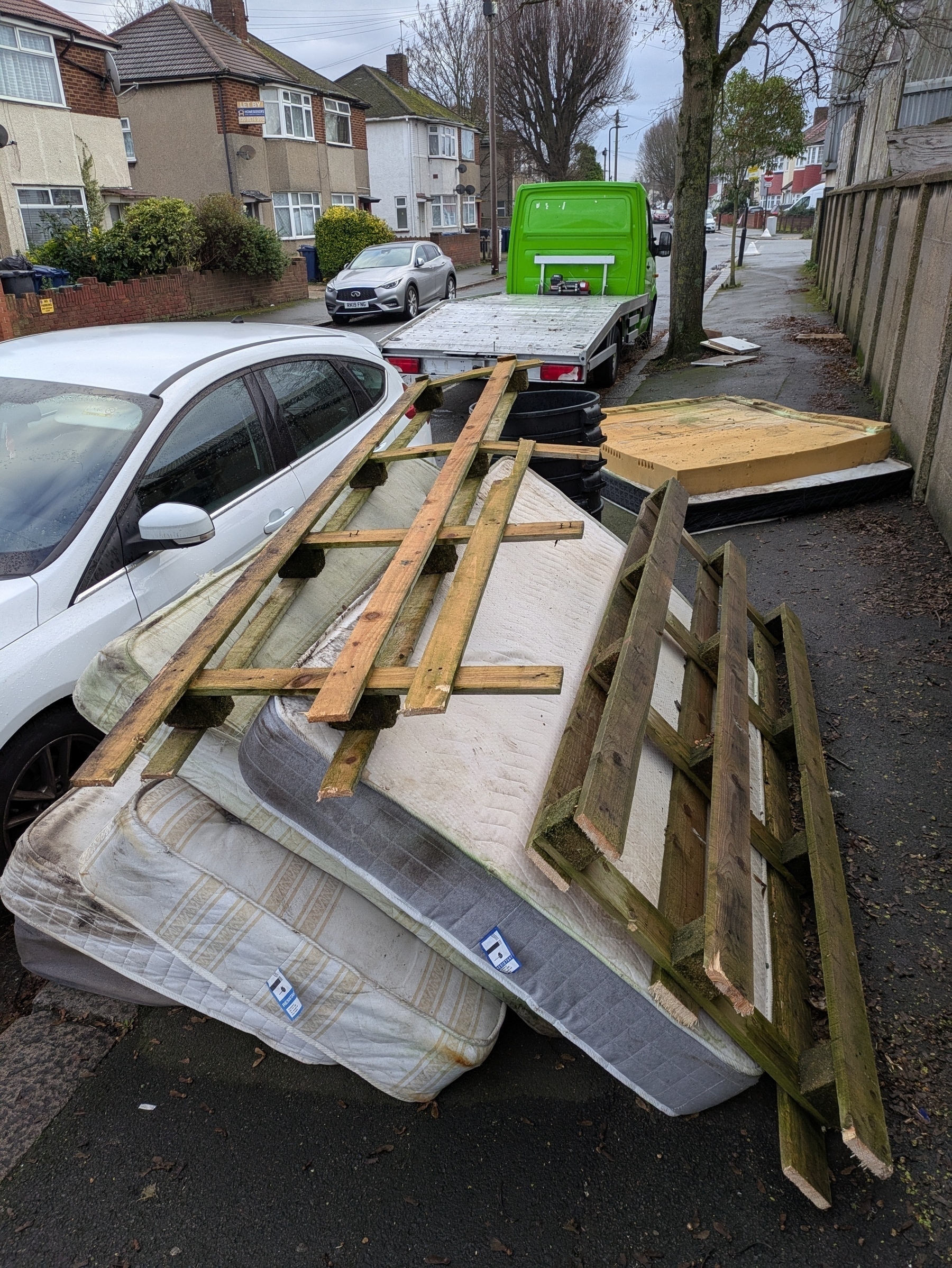 Auto-generated description: A pile of mattresses, wooden pallets, and other debris is stacked on a sidewalk next to a white car and a trailer.