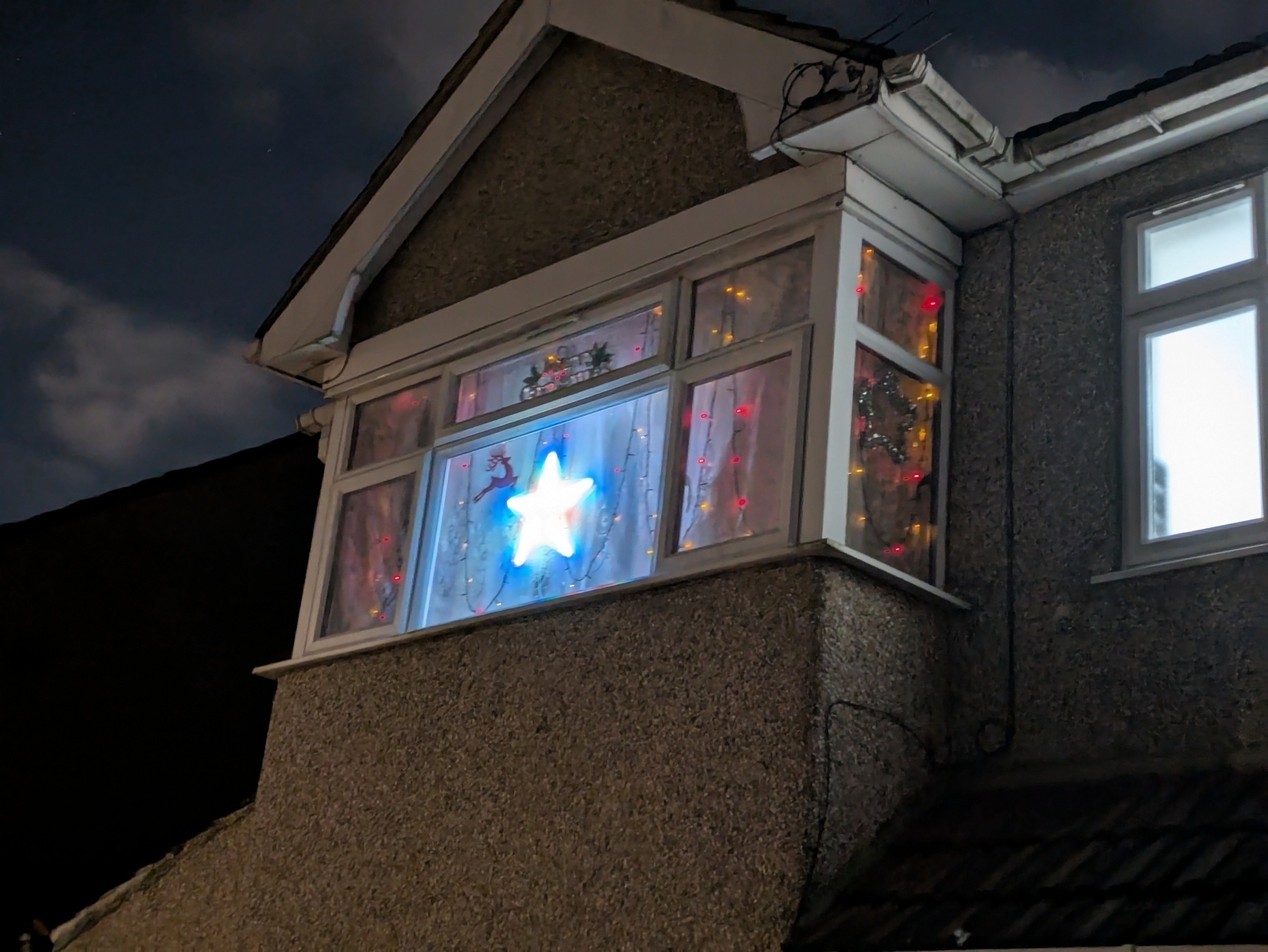 A brightly lit star decoration is displayed in the window of an upper floor of a house at night, surrounded by other festive lights and decor.