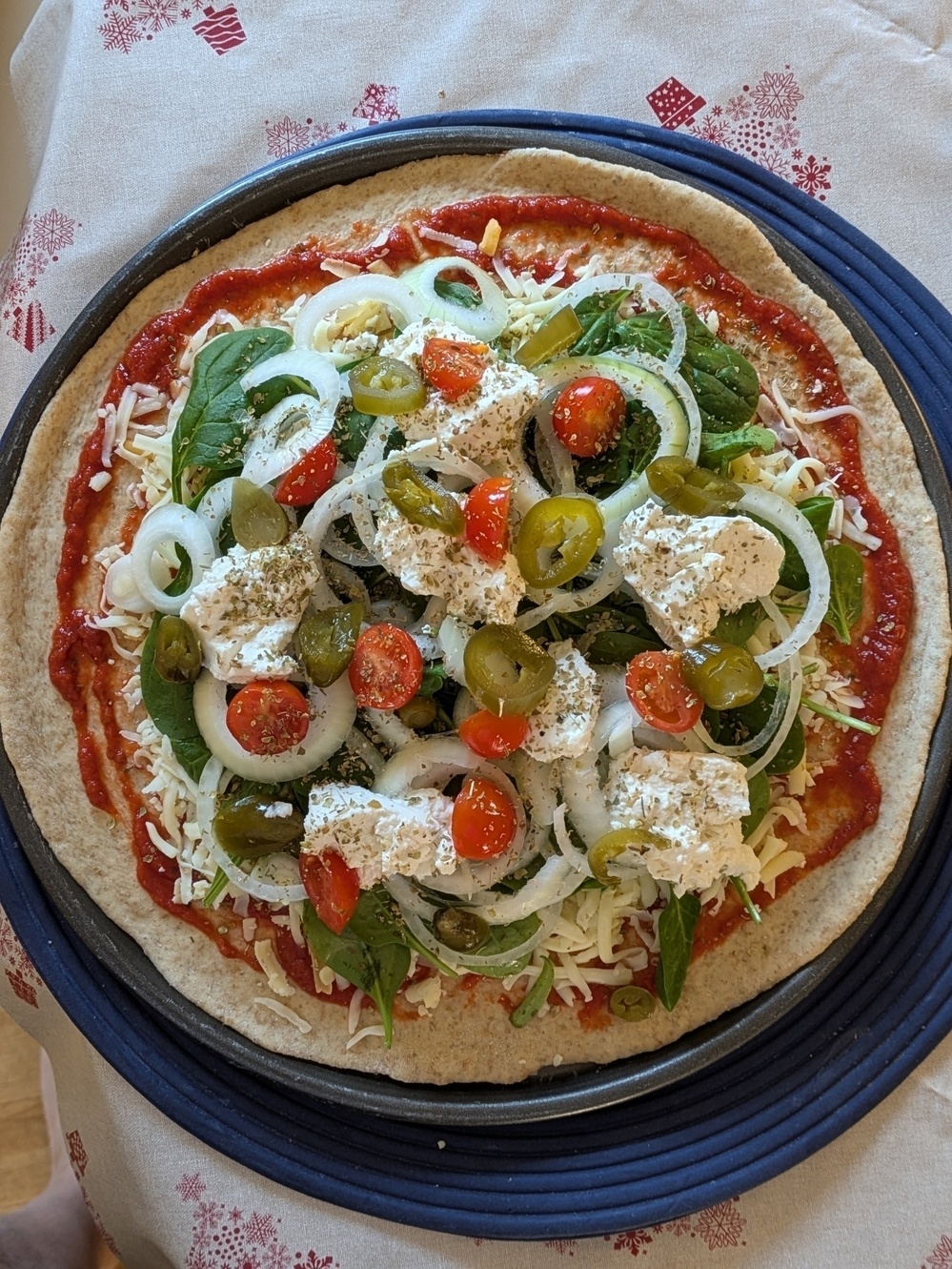 A pizza topped with onions, cherry tomatoes, cheese, spinach, jalapeños, and herbs is ready for baking.