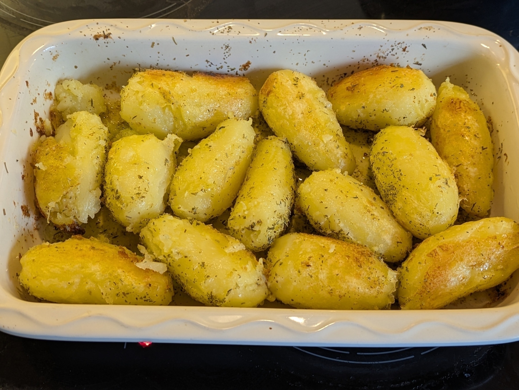 A rectangular beige ceramic baking dish on a black stove top filled with roasting whole potatoes garnished with herbs de provence