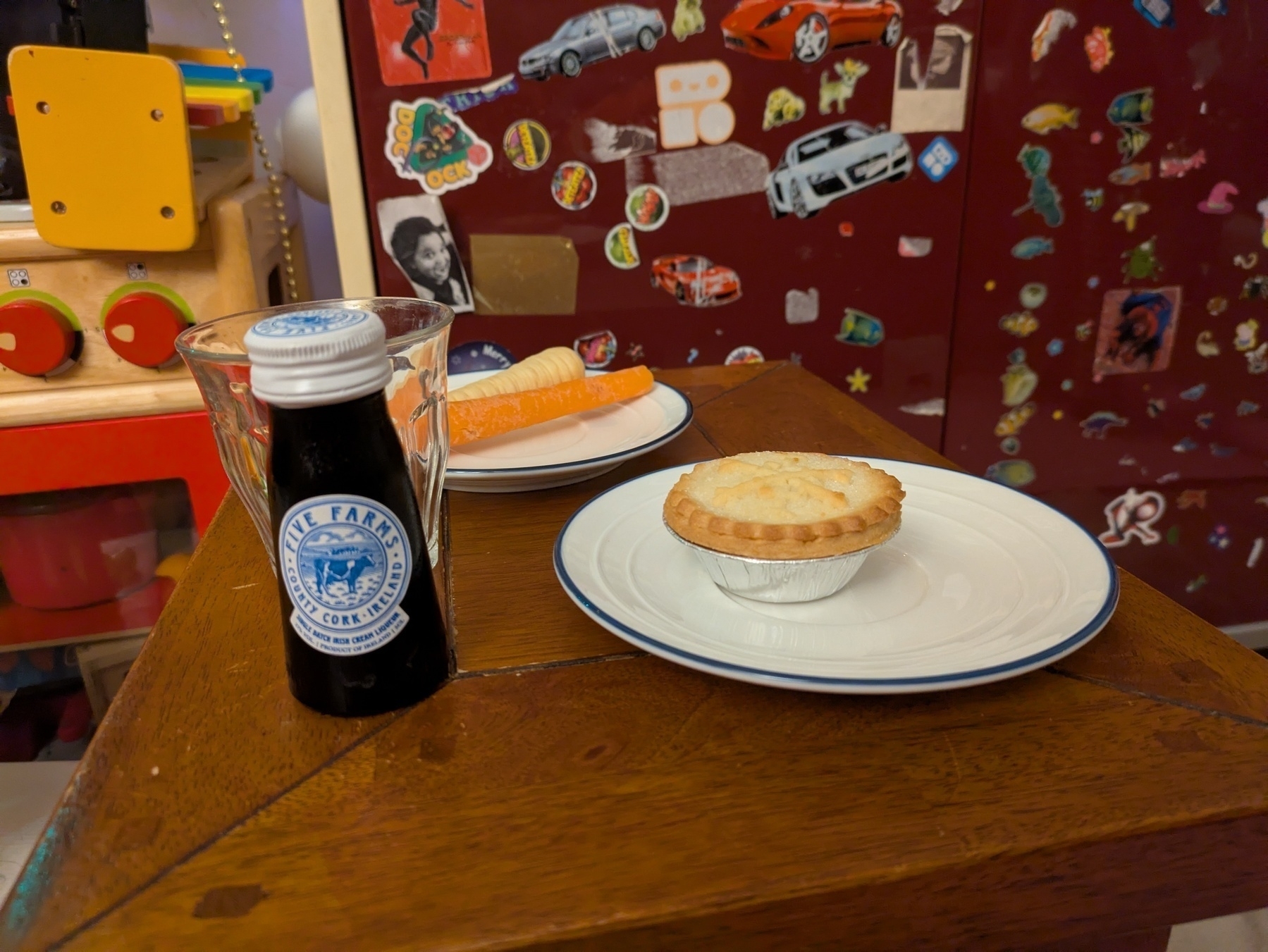 A small mahogany table holds a bottle of Five Farms Irish Cream Liqueur, a mince pie on a saucer and a carrot and a parsnip on another saucer, in front of a toy oven and a dark red cupboard adorned with stickers.