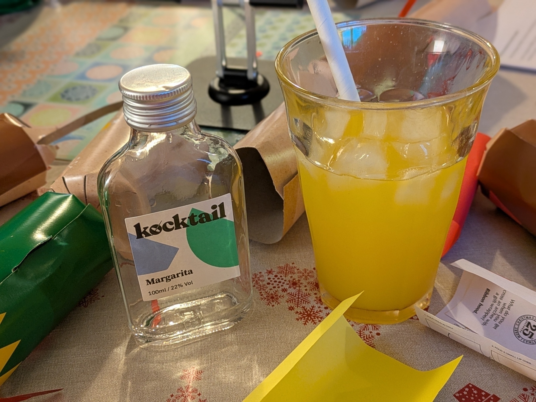 A glass of orange cocktail with ice and a straw is placed next to a small bottle labeled "kocktail Margarita" amid holiday-themed decorations.