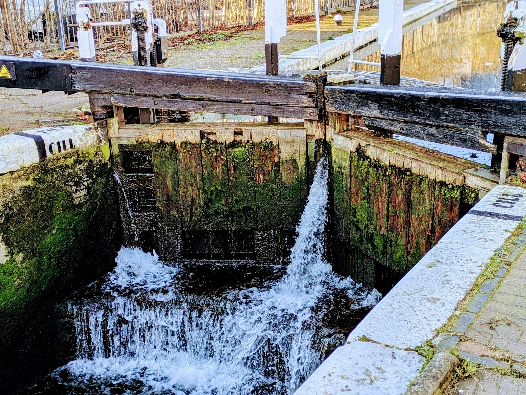 A canal lock is seen with water flowing through the partially open gate, surrounded by weathered wood and stone.