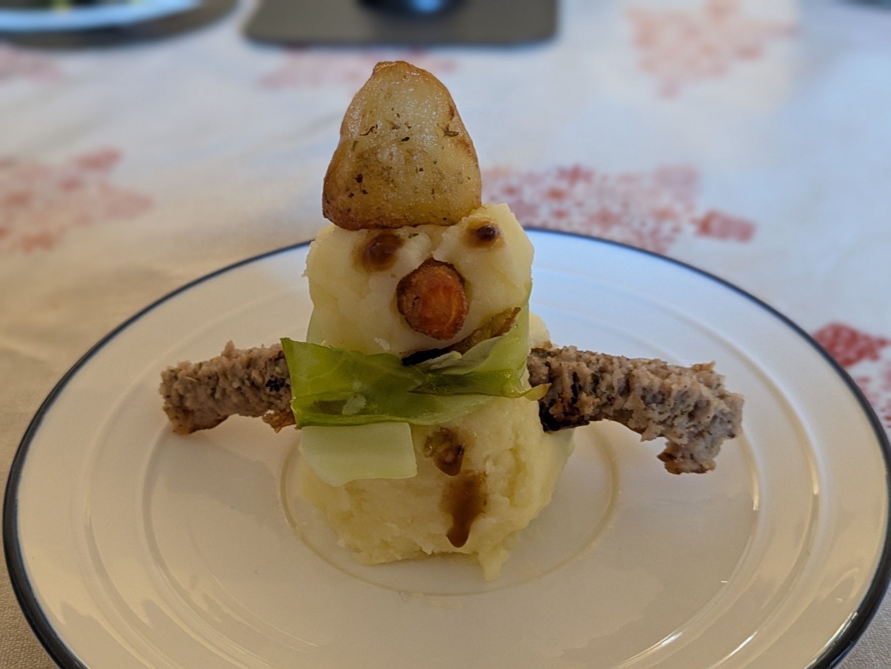 A snowman-shaped sculpture is made from mashed potatoes with a potato hat, carrot nose, gravy eyes, and meat for arms, served on a plate.