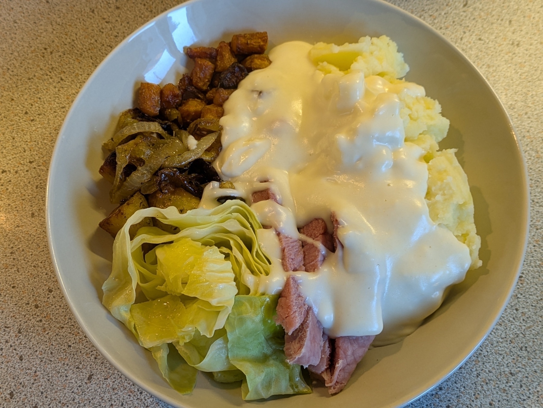 A bowl filled with mashed potatoes, cabbage, sliced meat topped with a creamy sauce, and a side of roasted vegetables.