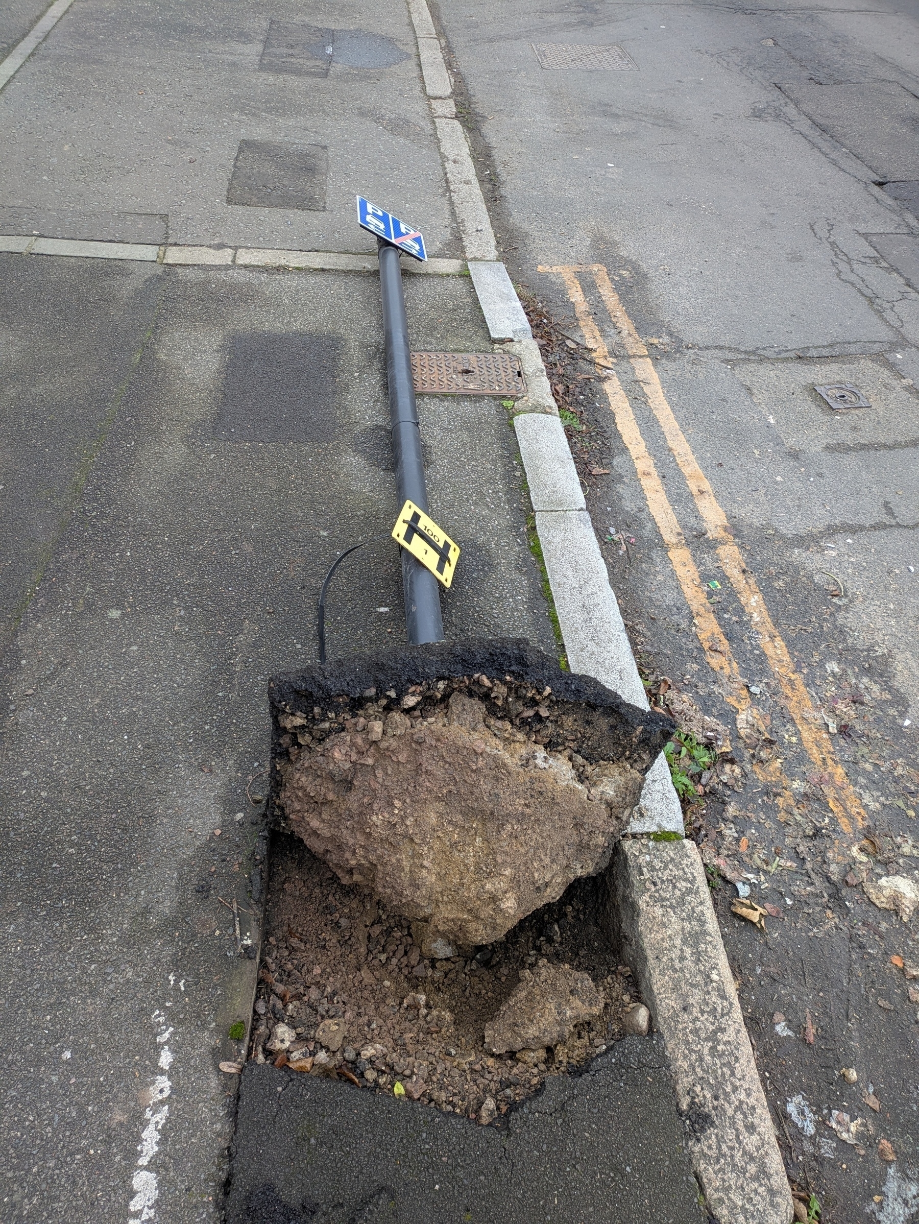 A fallen street sign lies uprooted on the sidewalk, exposing the base and surrounding pavement.