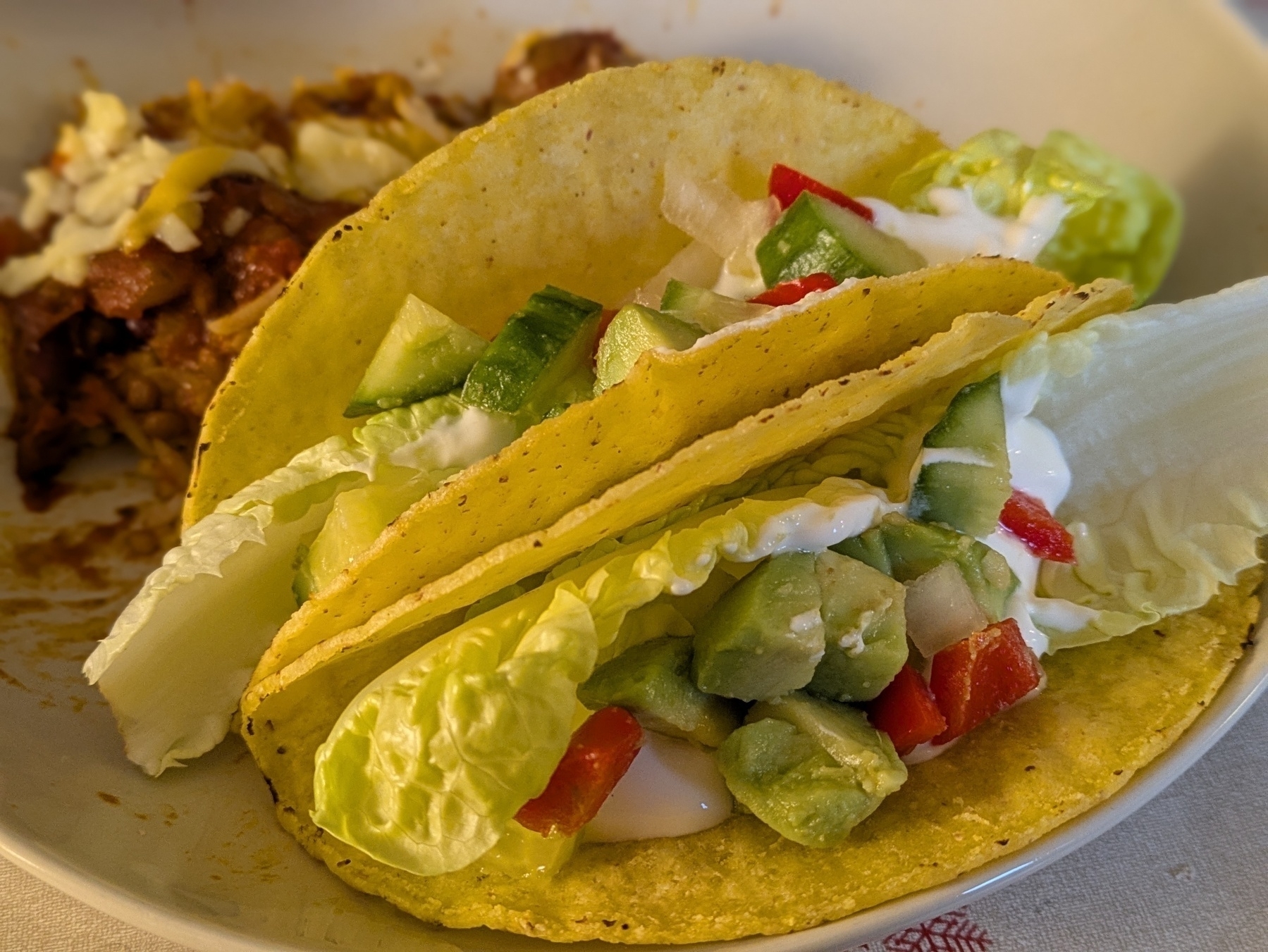 Two tacos filled with lettuce, avocado, cucumber, and red bell pepper sit on a plate next to a serving of chilli sans carne topped with cheese.