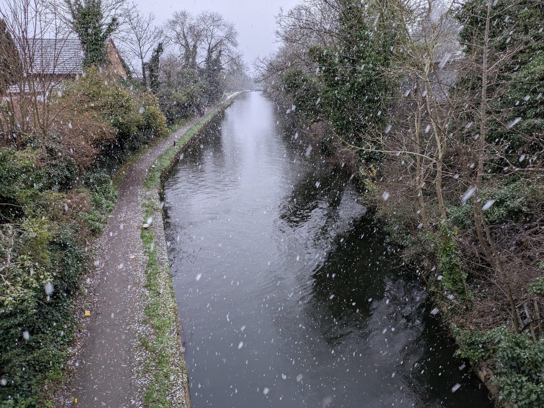 A narrow canal is flanked by trees and a path, with snow falling gently over the scene.