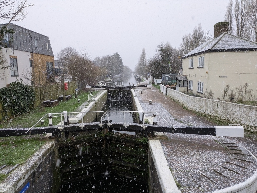 A snowy canal lock is flanked by buildings and trees on a wintry day.