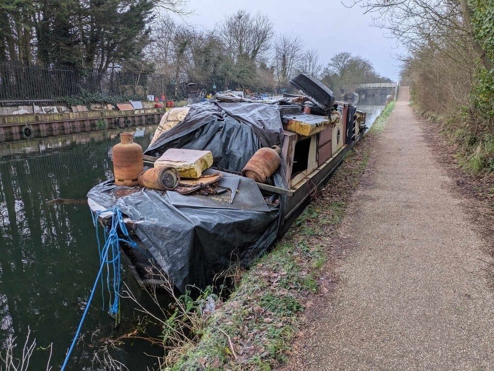 A narrow canal boat, covered with tarps and various items, is moored alongside a path surrounded by trees and vegetation.