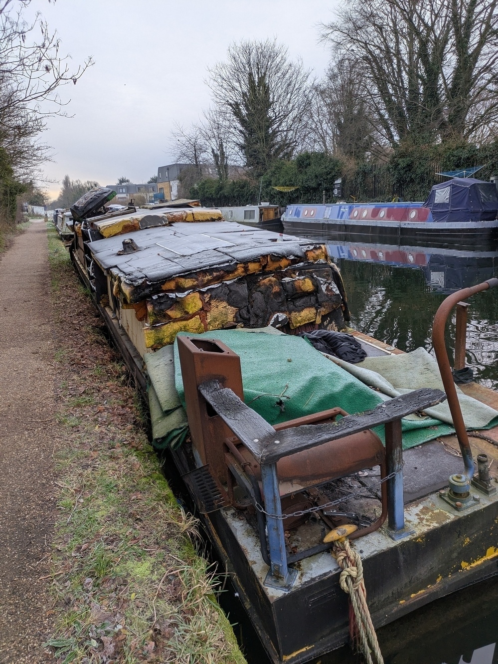 A damaged and partially covered narrowboat is moored along a canal surrounded by other boats and a pathway.