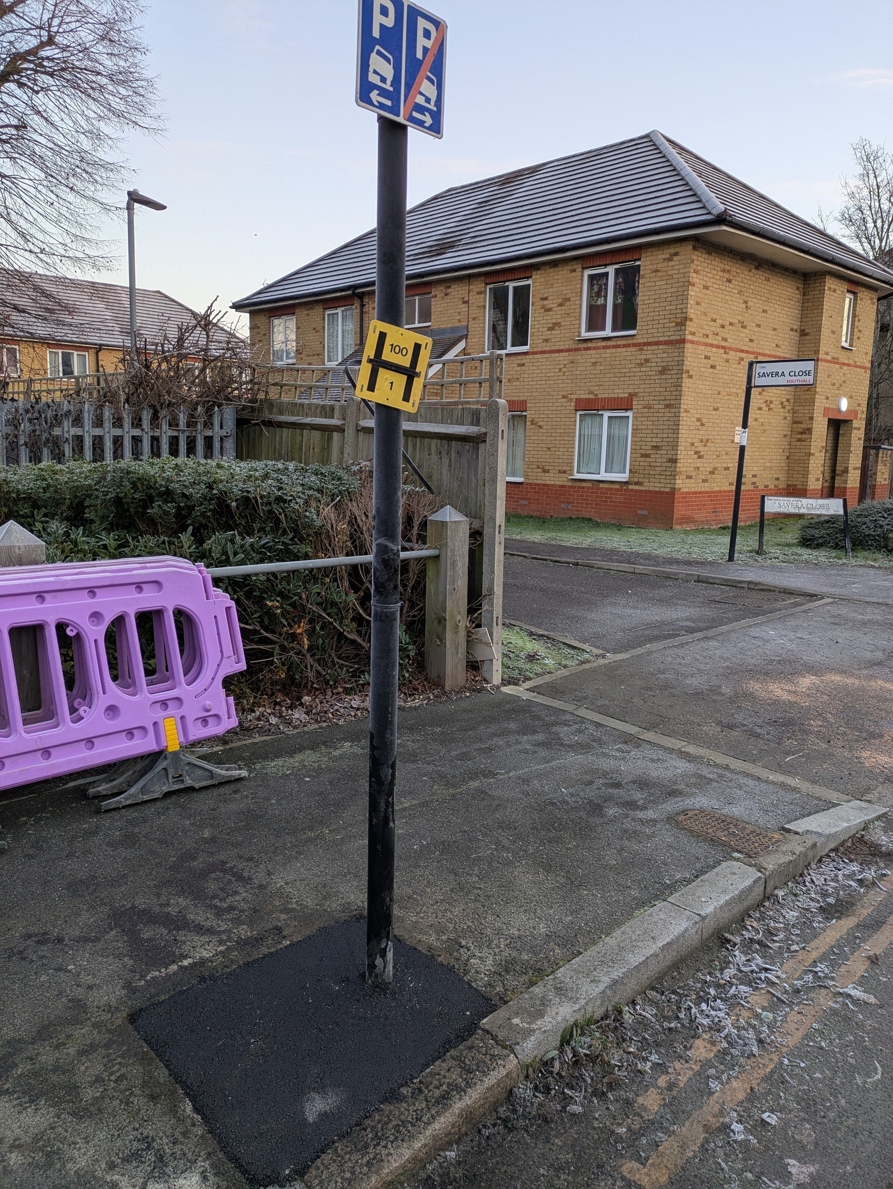 A parking sign is attached to a pole that's been newly installed on a sidewalk next to a purple barricade and a residential building.