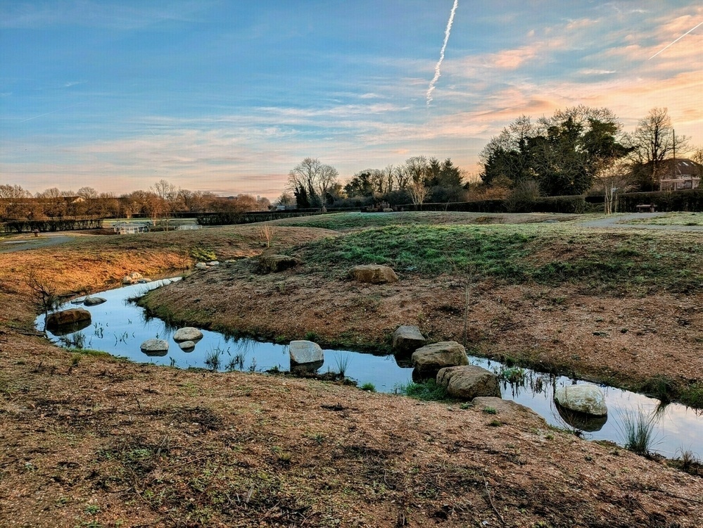 A small creek with stepping stones winds through a grassy landscape under a sky with streaks of clouds.