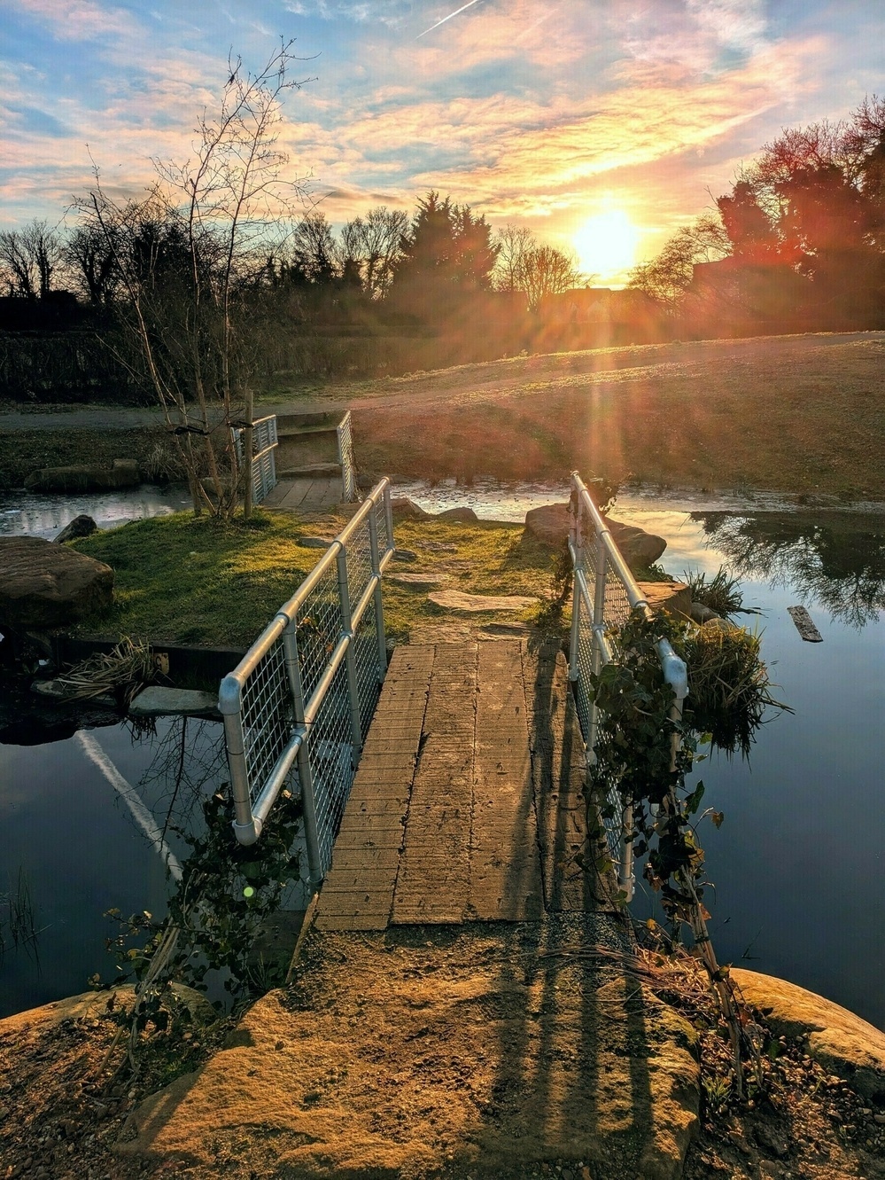 A small wooden bridge crosses over a tranquil creek, with a brilliant sunset in the background.