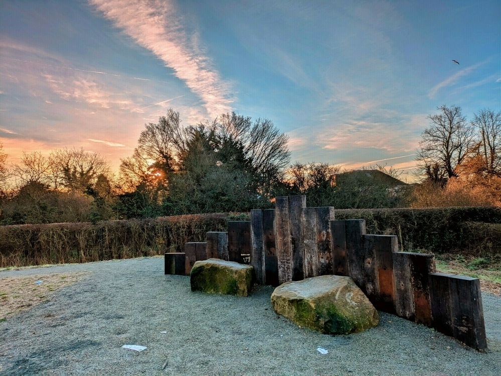 A tranquil outdoor scene features a row of wooden pillars next to large stones, surrounded by bare trees and a vivid sky at sunset.