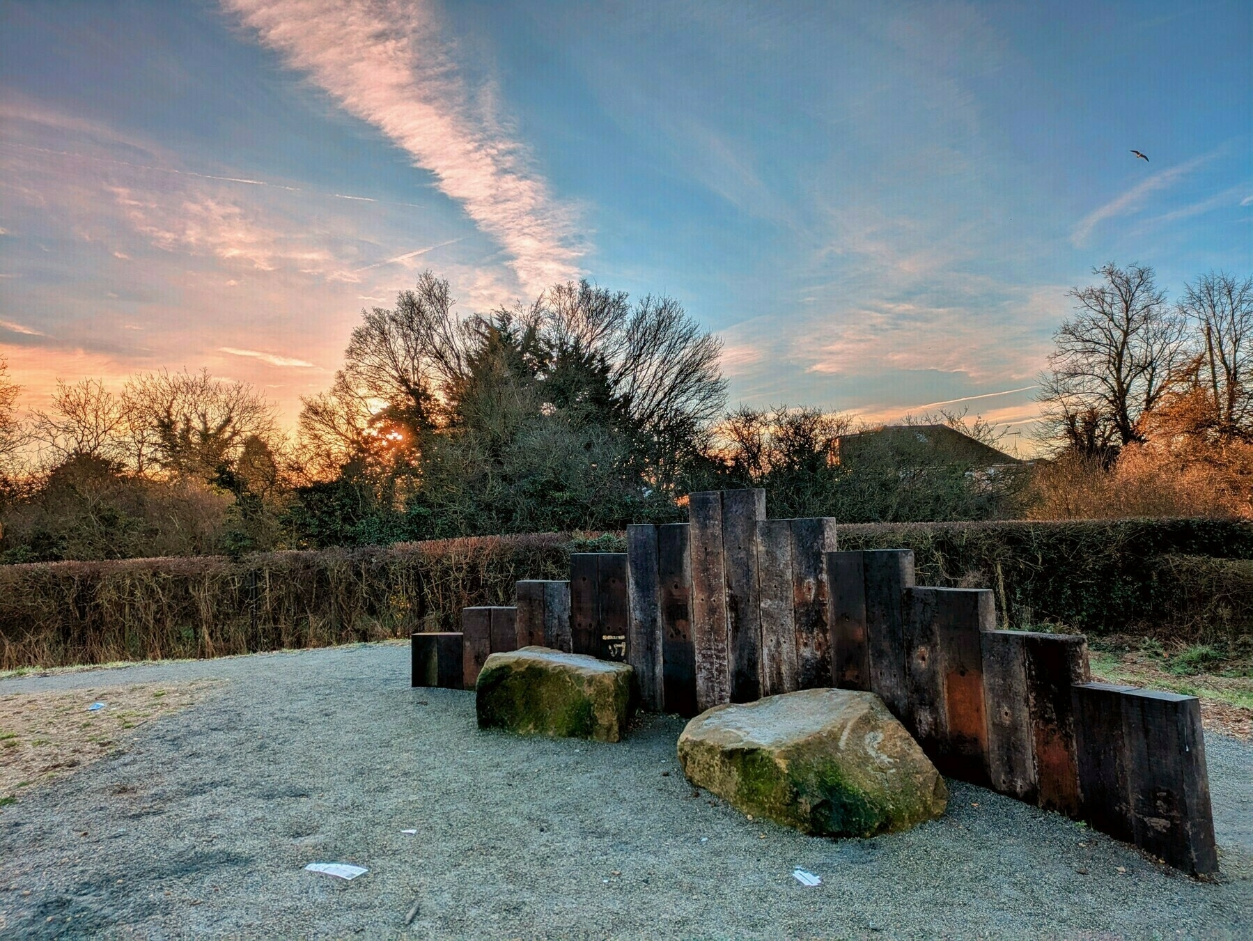 A tranquil outdoor scene features a row of wooden pillars next to large stones, surrounded by bare trees and a vivid sky at sunrise.