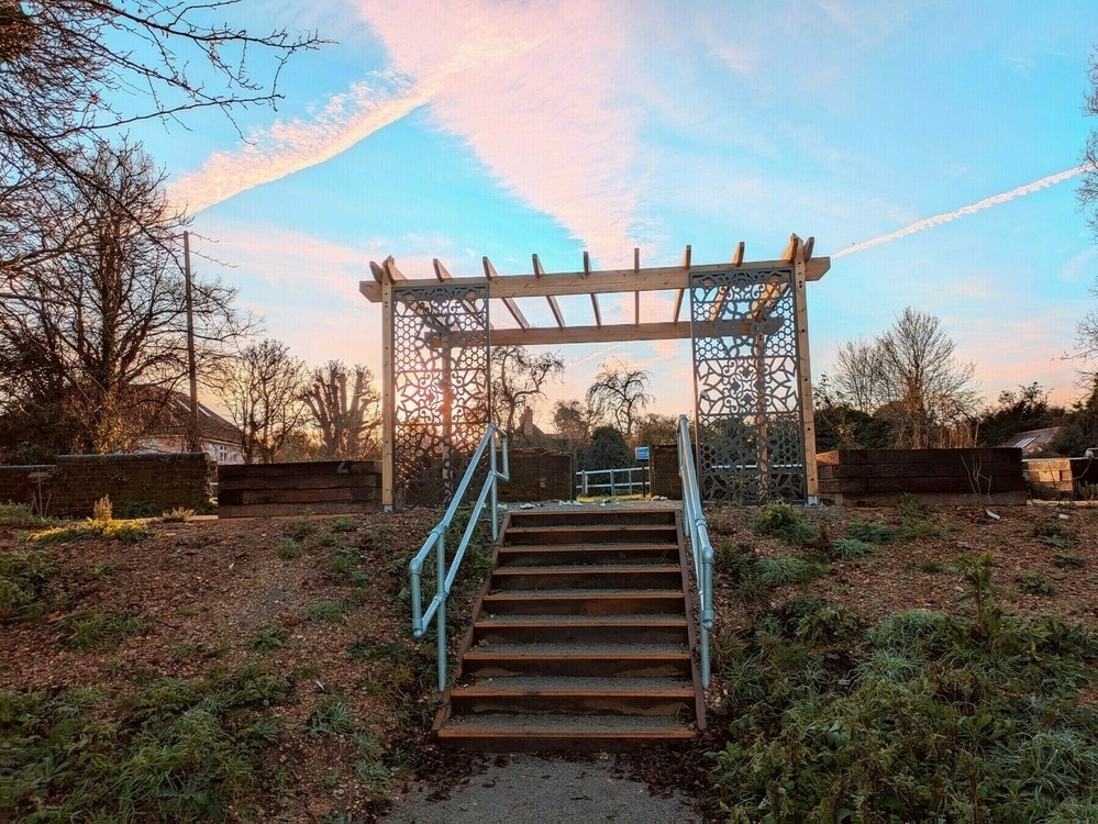 A wooden pergola stands at the top of a set of stairs surrounded by trees and a blue sky with clouds.