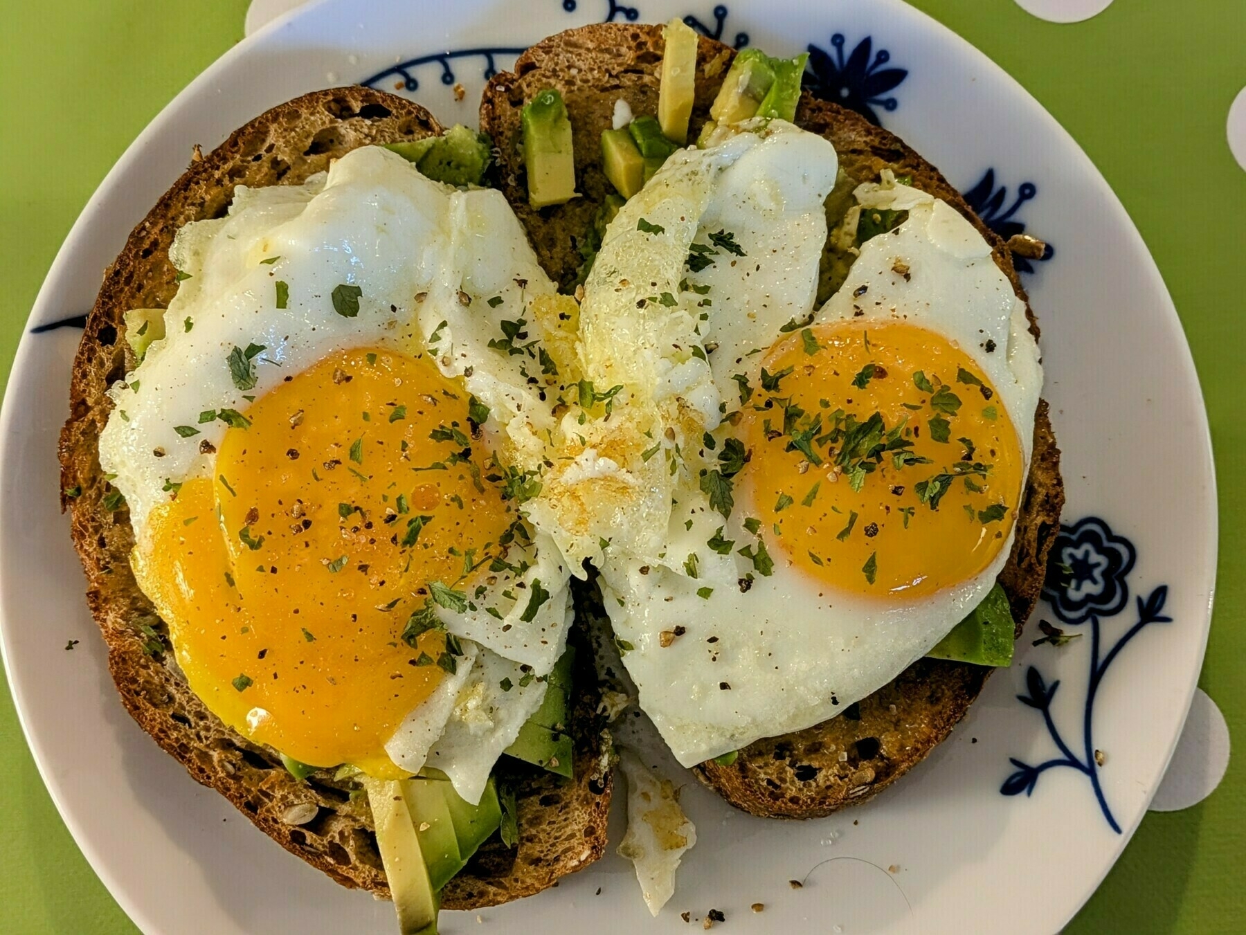 Two slices of toasted seeded multigrain bloomer bread topped with chopped avocado and two fried eggs on a white plate patterned with navy blue swirls on a lime green tablecloth with white polka dots.